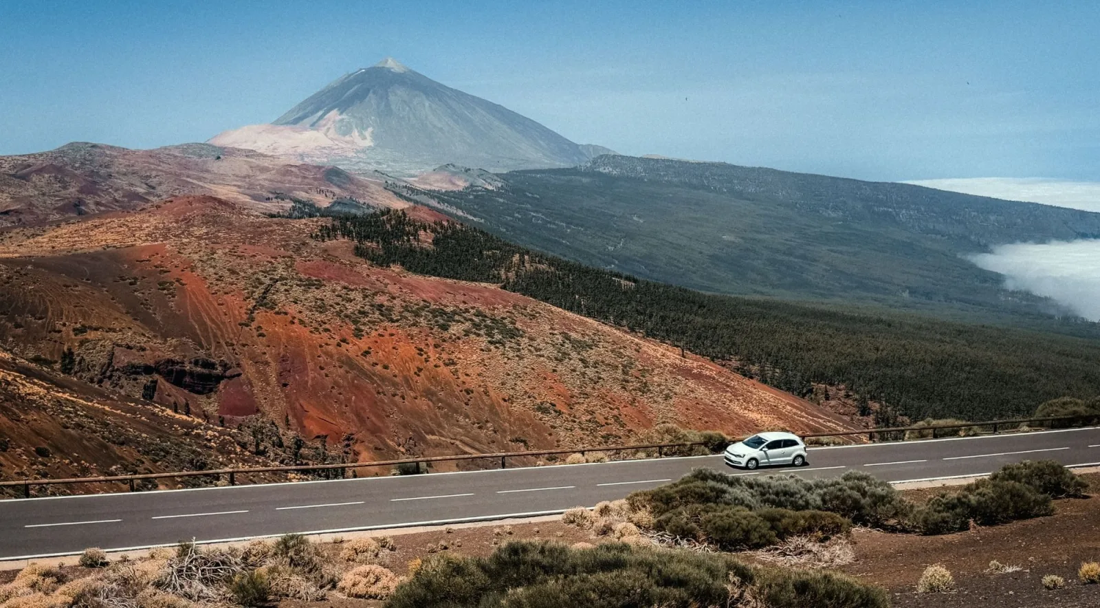 The cone of Mount Teide above red volcanic hills, with a car on the mountain road and a sea of cloud to the right