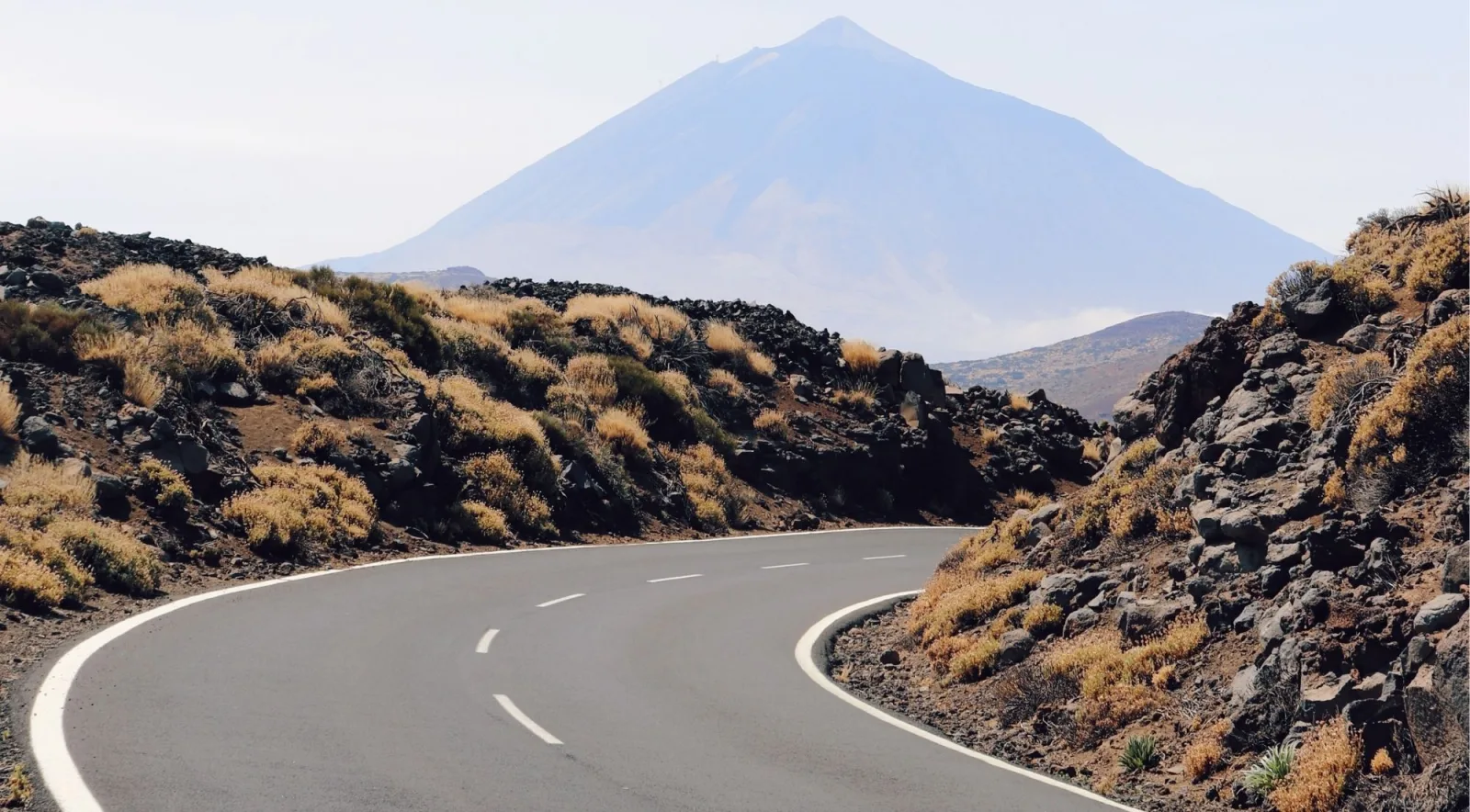 A curve in the park road with dry grasses and black lava boulders, Mount Teide hazy in the distance