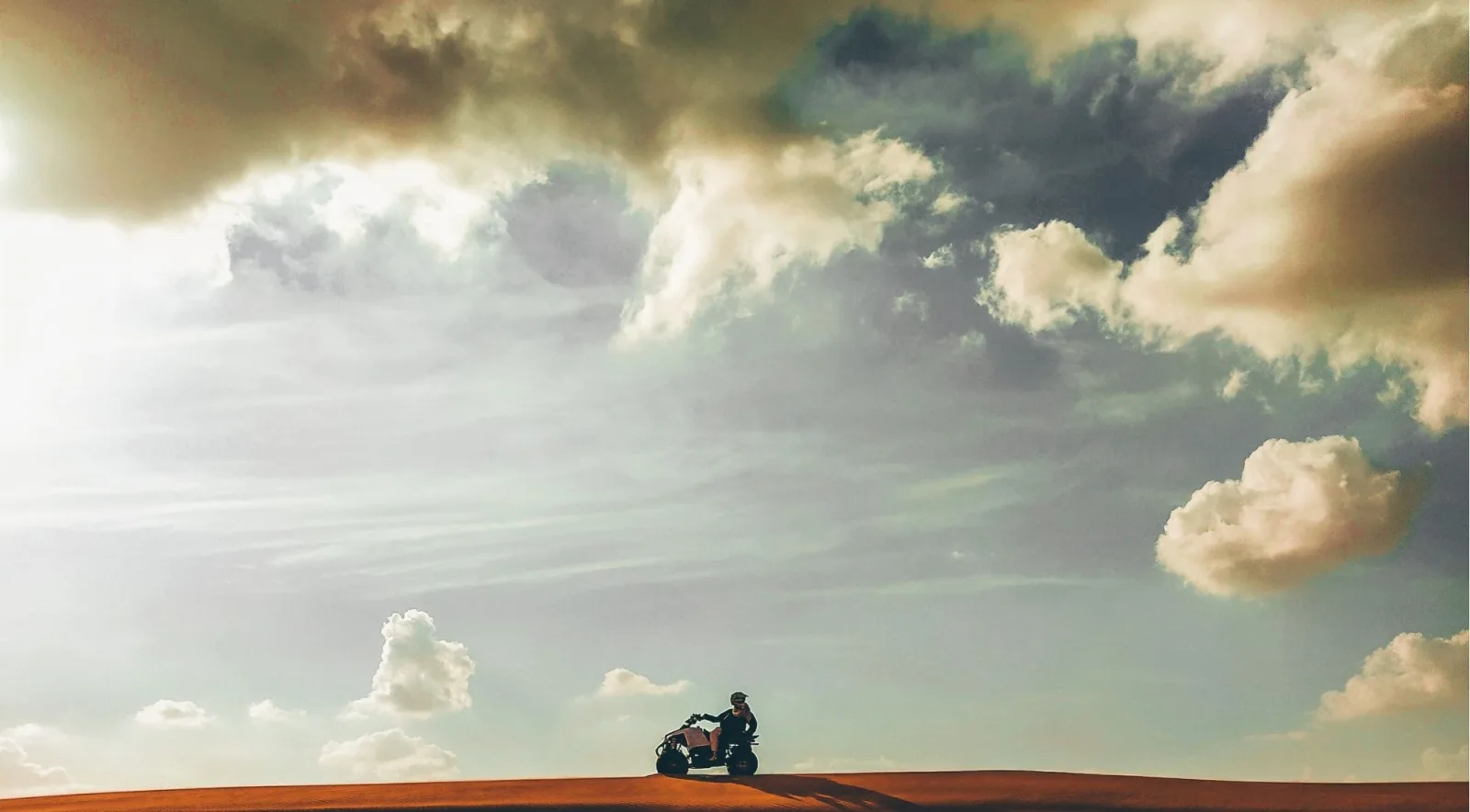 A lone quad rider silhouetted on a ridge under a dramatic cloudy sky